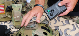 An operator's hands are shown touching a strobe light on a camouflage helmet while holding the black PIM-701 remote control unit ready to transmit new light sequences.