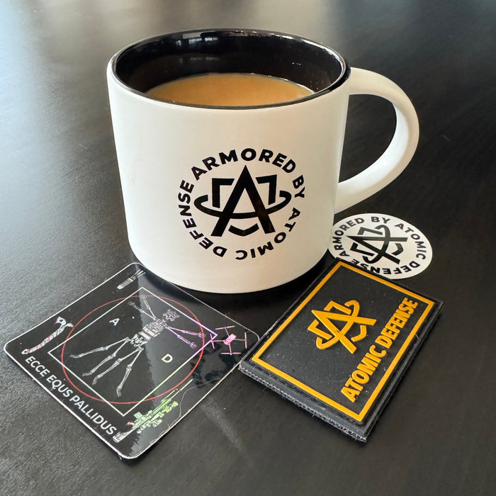 A detailed profile shot of the Atomic Defense coffee mug surrounded by various complimentary freebies included in every order, such as branded tactical patches and colorful stickers scattered neatly on the table.