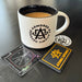A detailed profile shot of the Atomic Defense coffee mug surrounded by various complimentary freebies included in every order, such as branded tactical patches and colorful stickers scattered neatly on the table.