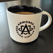 A clean side profile shot of a minimalist black and white Atomic Defense 16oz coffee mug, featuring the brand's logo prominently displayed in the foreground against a smooth surface.