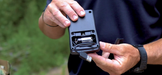 A person holds the black programming module with the back cover removed, revealing the internal storage for the USB cable and the Single Replaceable CR123 battery that powers the unit.