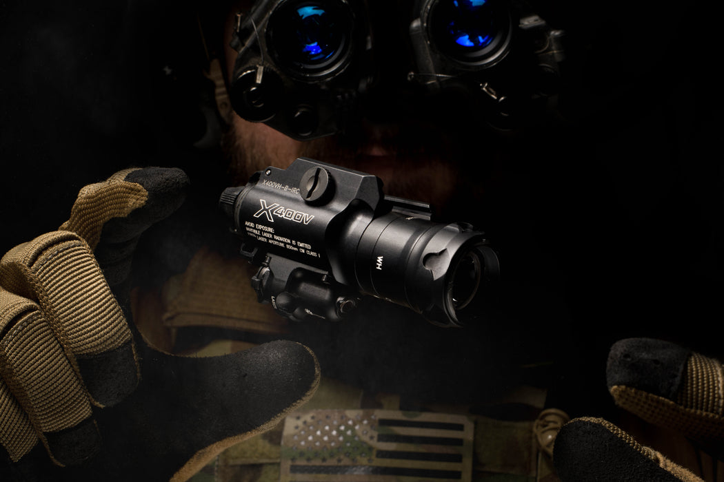 A dramatic profile shot of a tactical operator wearing night vision goggles (NODs), holding the X400VH Vampire in their open palm to showcase the unit's compact size and the integrated infrared laser emitter.