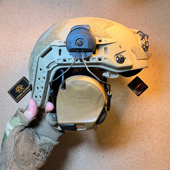 A gloved hand holds a tan high-cut helmet equipped with a coyote brown 3M Peltor ComTac tactical headset mounted directly to the side ARC rails.