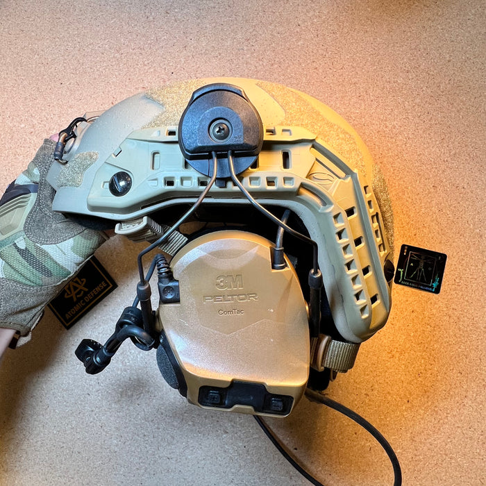 A side angle of a tan high-cut ballistic helmet showing the helmet rail attachment system firmly securing a coyote brown Peltor ComTac VIII hearing protection earcup.