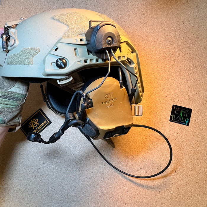 A close-up view of a tan high-cut helmet featuring a rail-mounted 3M Peltor ComTac system with the noise-canceling boom microphone extended downward for clear battle comms.