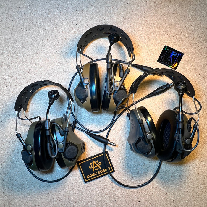 Three Peltor ComTac VIII communication systems are arranged together on a textured surface, showcasing the available olive drab, coyote brown, and black color options next to an Atomic Defense patch.