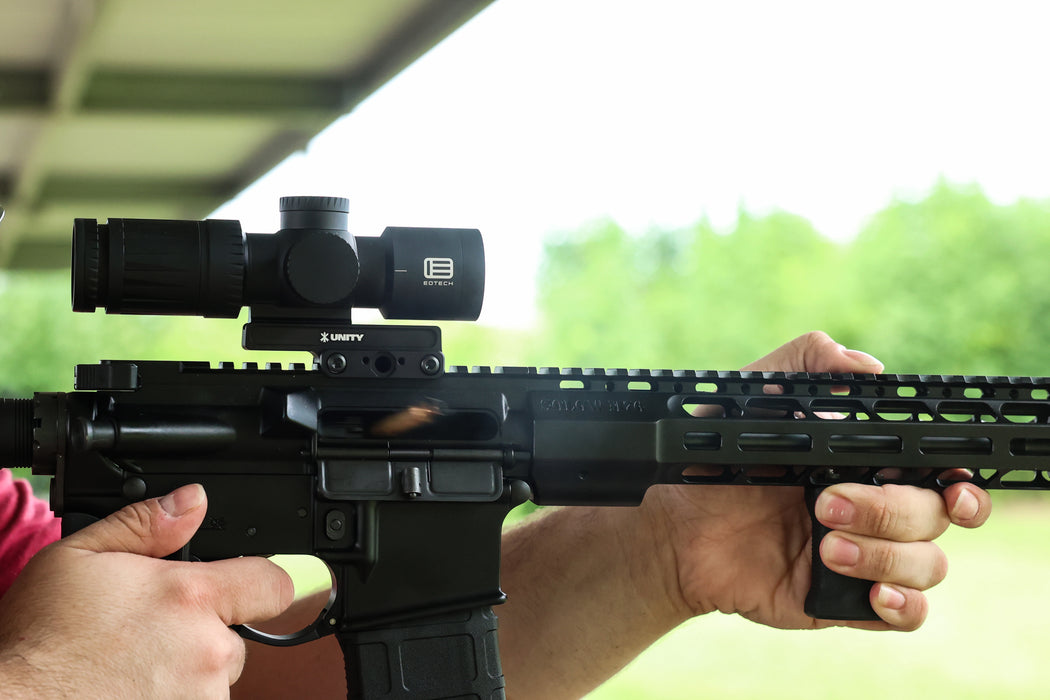A side profile action shot of a black Cobalt Kinetics AR-15 firing, with a spent casing expelling from the chamber due to recoil. The rifle is equipped with a black Unity FAST Kickr mount, demonstrating its ability to maintain a steady 1.54-inch sight picture under heavy vibration.